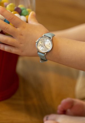 Montre en argent avec de grands chiffres sur un cadran blanc, bracelet en cuir bleu clair, portée sur le poignet d'un enfant, avec un pot de bonbons colorés en arrière-plan.