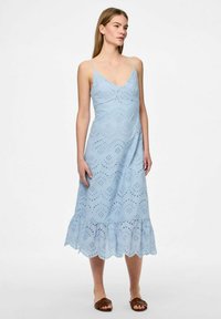 Gaiši zila kokvilnas kleita ar V-veida kakla izgriezumu, mežģīņu acs rakstu un villīgiem izgriezumiem apakšmalā. Dizains ietver plānas siksnas un piegulošu siluetu.