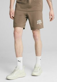 Pantalones cortos de algodón marrón con el logo blanco "PUMA sports club" en la pierna, combinados con calcetines deportivos blancos y zapatillas crema. Diseño minimalista.