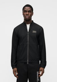 Melnā jaciņa ar rāvējslēdzēju, reljefu rakstu, stāvošu apkakli un izšūtu emblēmu. Kombinēta ar piemērotām biksēm ar baltu zīmolu.