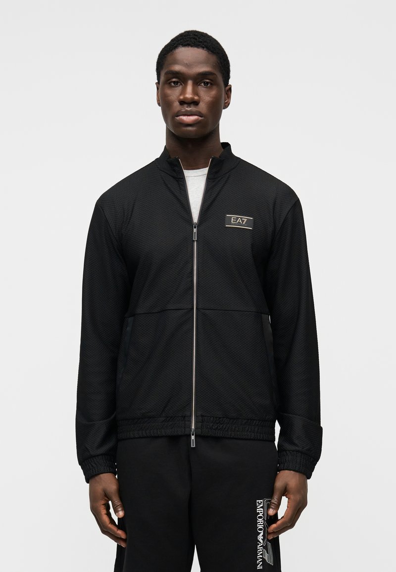 Melnā jaciņa ar rāvējslēdzēju, reljefu rakstu, stāvošu apkakli un izšūtu emblēmu. Kombinēta ar piemērotām biksēm ar baltu zīmolu.