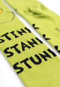 Calzini verdi con una stampa di un personaggio dei cartoni animati sorridente e testo nero in grassetto che recita "STINK," "STANK," e "STUNK." Tessuto in maglia morbido.
