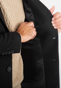 Manteau extérieur en laine noire avec un intérieur en satin. Le porteur ajuste une poche, mettant en valeur un pull beige texturé en dessous.