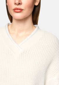 Nahaufnahme einer Frau, die einen weißen, strukturierten Strickpullover mit V-Ausschnitt und weißem Innenkragen trägt. Zu sehen sind ihre Lippen, ihr Hals und ein Teil ihres Haares.