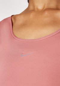 Ljusrosa träningslinne med rundad halsringning och tunna axelband, försett med en liten, präglad grå Nike-logotyp på bröstet. Slät textur.