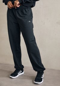 Melni sporta bikses ar elastīgu jostu, kurās ir diskrēts Adidas logo. Vienotas ar melniem un baltiem sporta apaviem, uz neitrāla fona.