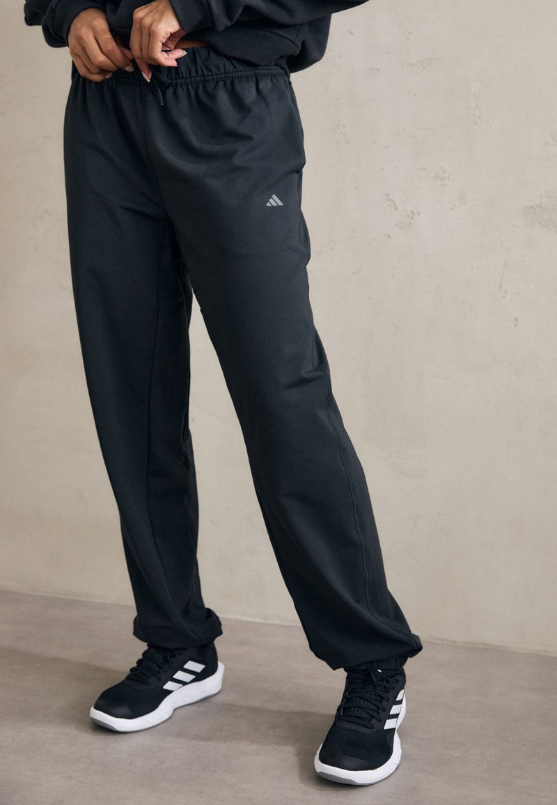 Melni sporta bikses ar elastīgu jostu, kurās ir diskrēts Adidas logo. Vienotas ar melniem un baltiem sporta apaviem, uz neitrāla fona.
