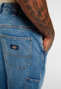 Light blue denim pants with a back pocket featuring a “Dickies” logo patch. Orange stitching and a classic fit design with a textured finish.