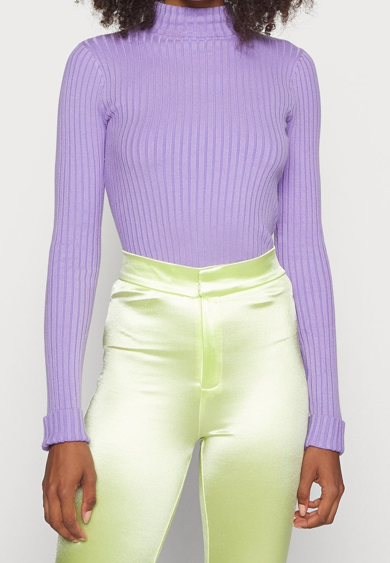 Femme portant un haut côtelé à manches longues lavande rentré dans un pantalon taille haute vert citron brillant, debout devant un fond uni.