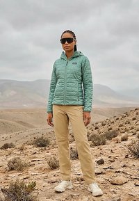 Žena u tirkiznoj prošivenoj jakni, bež hlačama, bijelim cipelama i velikim sunčanim naočalama stoji u kamenitoj pustinjskog krajoliku pod oblačnim nebom.