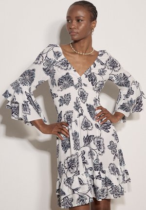 Kobieta w białej sukience z czarnym kwiatowym wzorem, dekoltem w serek oraz warstwowymi rozkloszowanymi rękawami, pozująca z rękami na biodrach na jednolitym tle.