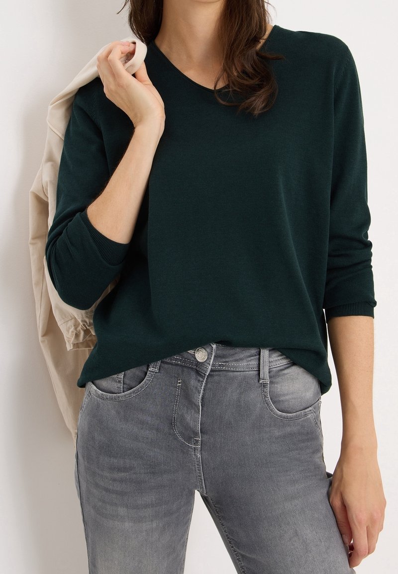 Femme portant un haut à manches longues vert foncé et un jean gris, tenant une veste beige sur son épaule, sur un fond uni.