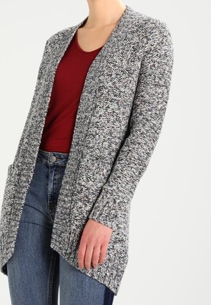 Femme portant un cardigan en maille gris avec des poches devant sur une chemise rouge et un jean bleu, montrée du cou aux hanches sur un fond uni.