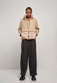 Beige cropped jacket met glanzend beige puffer onderkant, rits aan de voorkant, hoge kraag; gecombineerd met donkergrijze broek met wijde pijpen en zwarte platform schoenen.
