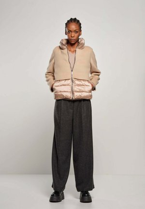Beige cropped jacket met glanzend beige puffer onderkant, rits aan de voorkant, hoge kraag; gecombineerd met donkergrijze broek met wijde pijpen en zwarte platform schoenen.
