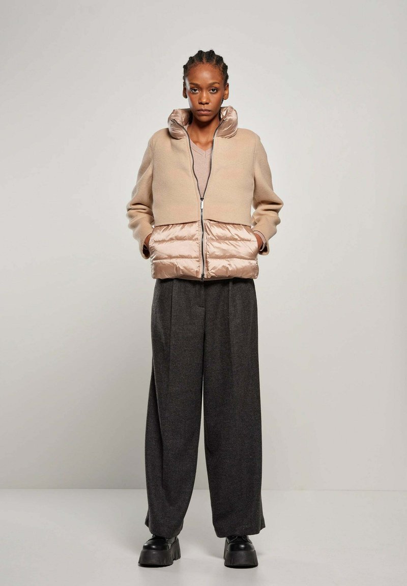 Beige cropped jacket met glanzend beige puffer onderkant, rits aan de voorkant, hoge kraag; gecombineerd met donkergrijze broek met wijde pijpen en zwarte platform schoenen.