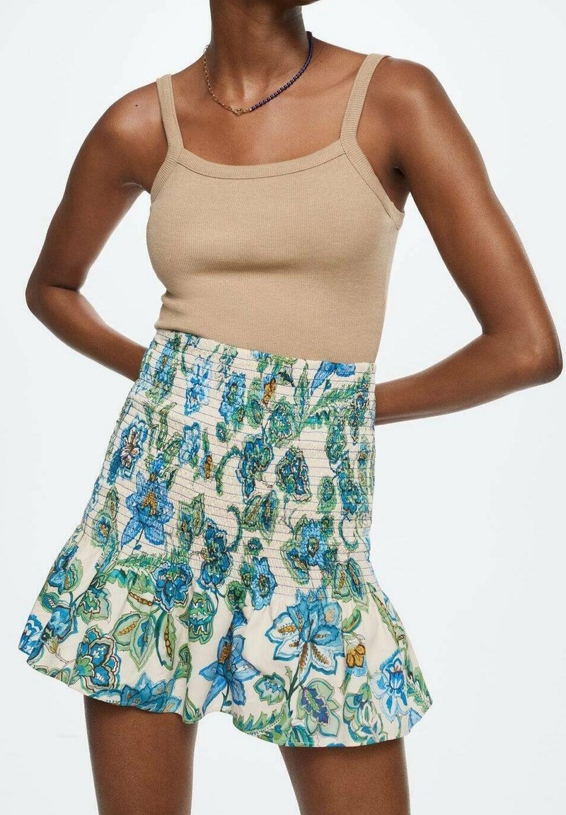 Femme portant un haut beige sans manches et une jupe taille haute bleu et vert à motif floral avec un ourlet à volants.