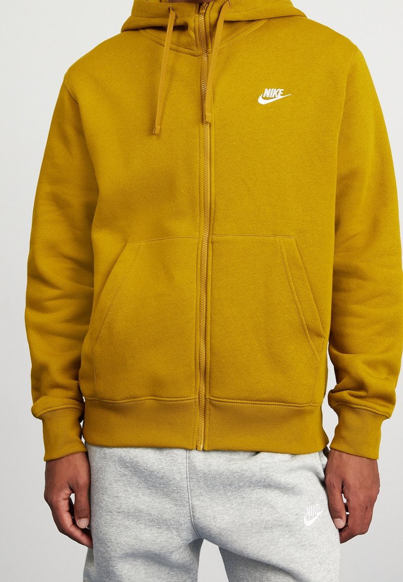 Sudadera amarilla con cremallera y cordones, bolsillos tipo canguro y logo blanco de Nike en el pecho. Tela suave y texturada, combinada con pantalones de chándal grises.