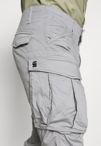 Szare spodnie robocze wykonane z fakturowanej tkaniny, z wieloma kieszeniami, przeszyciami oraz małym logo na tylnej kieszeni.