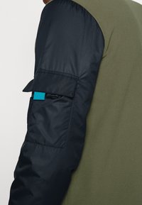 Olivově zelený fleec na jedné straně přechází do tmavě námořnického prošívaného rukávu s modrým akcentem na kapse. Viditelné funkční designové prvky.