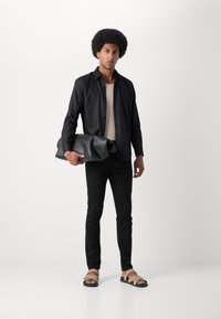 Musta pitkähihainen paita vaaleanraidallisen hihattoman topin päällä, mustat slim fit -housut ja ruskeat narusaappaat. Pitelee suurta harmaata pehmeästä nahasta valmistettua laukkua.