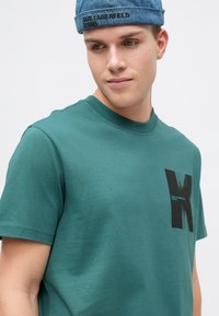 Zelené bavlněné tričko s krátkým rukávem, kulatým výstřihem a černým grafickým logem na levé straně hrudi; hladká textura. Džínová čepice s logem.