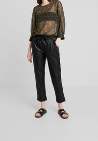 Läpinäkyvä musta kukkakuviollinen toppi kolmannelta neljännekseltä hihoilla, yhdistettynä tiukkoihin mustiin keinonahkahousuihin ja matalapohjaisiin mustiin sandaaleihin.