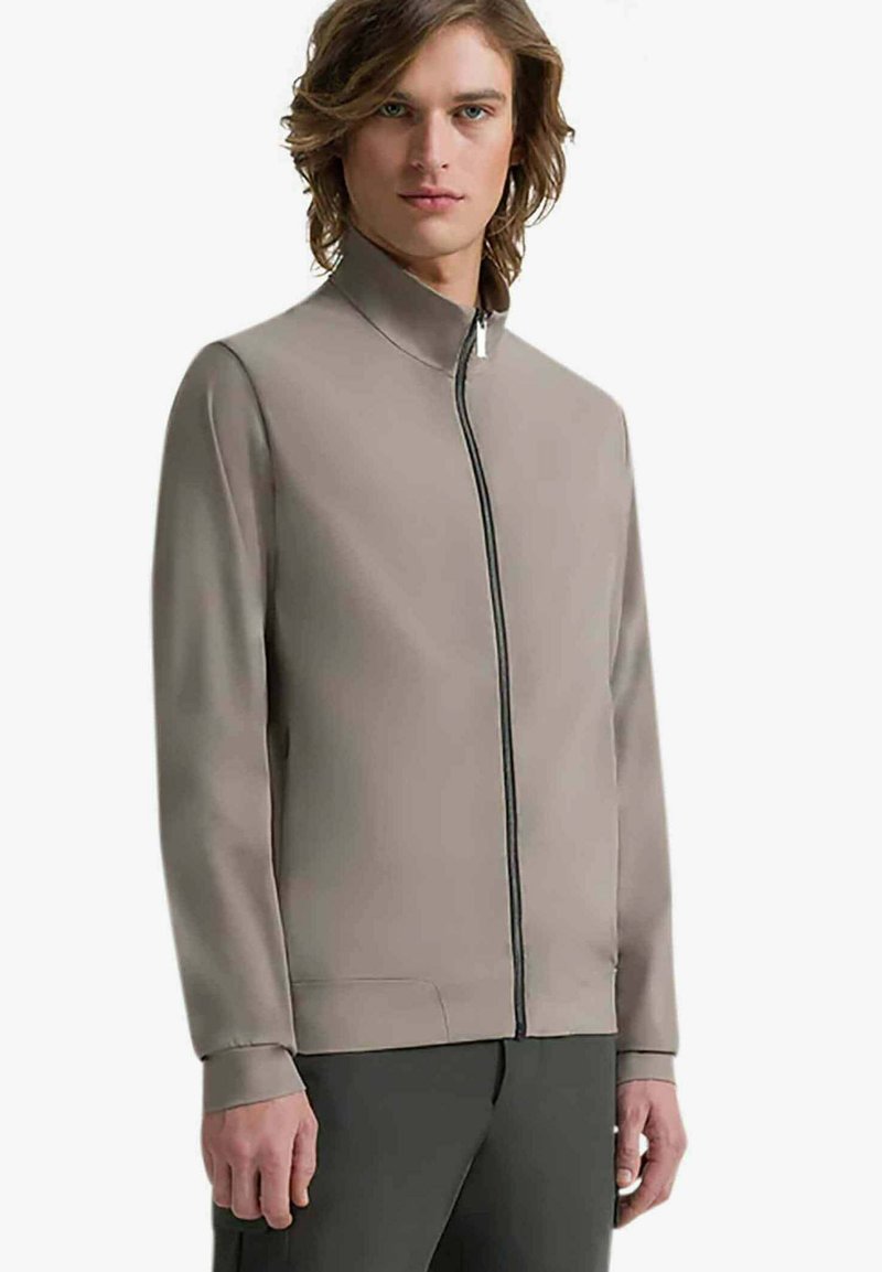 Giacca taupe chiaro con colletto alto, zip frontale e maniche lunghe. Caratterizzata da una texture liscia e un design minimalista.