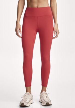 Woman wearing red high-waist leggings and white athletic shoes standing against a plain light background.