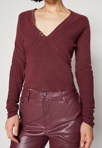 Bordowy prążkowany sweter z dzianiny z dekoltem w serek, ozdobiony srebrnymi elementami przy dekolcie. Na zdjęciu również dopasowane, błyszczące bordowe skórzane spodnie.