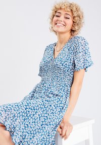 Robe florale bleue à manches courtes, corsage smocké et ourlet volanté, avec des fleurs blanches sur fond bleu foncé.