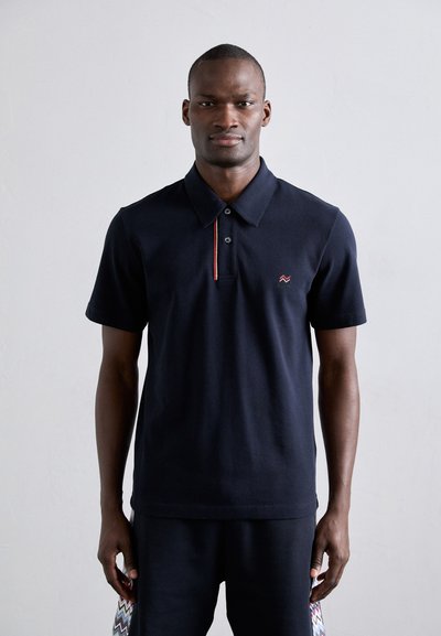 Marineblå polo shirt med krave, der har en rød accentstripe og logo, lavet af et struktureret stof, båret med mørke matchende bukser.