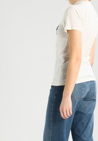 Bavlněné tričko v barvě přírodní bílé s kulatým výstřihem a krátkými rukávy. Spáváno s modrými džíny, které mají volný střih.