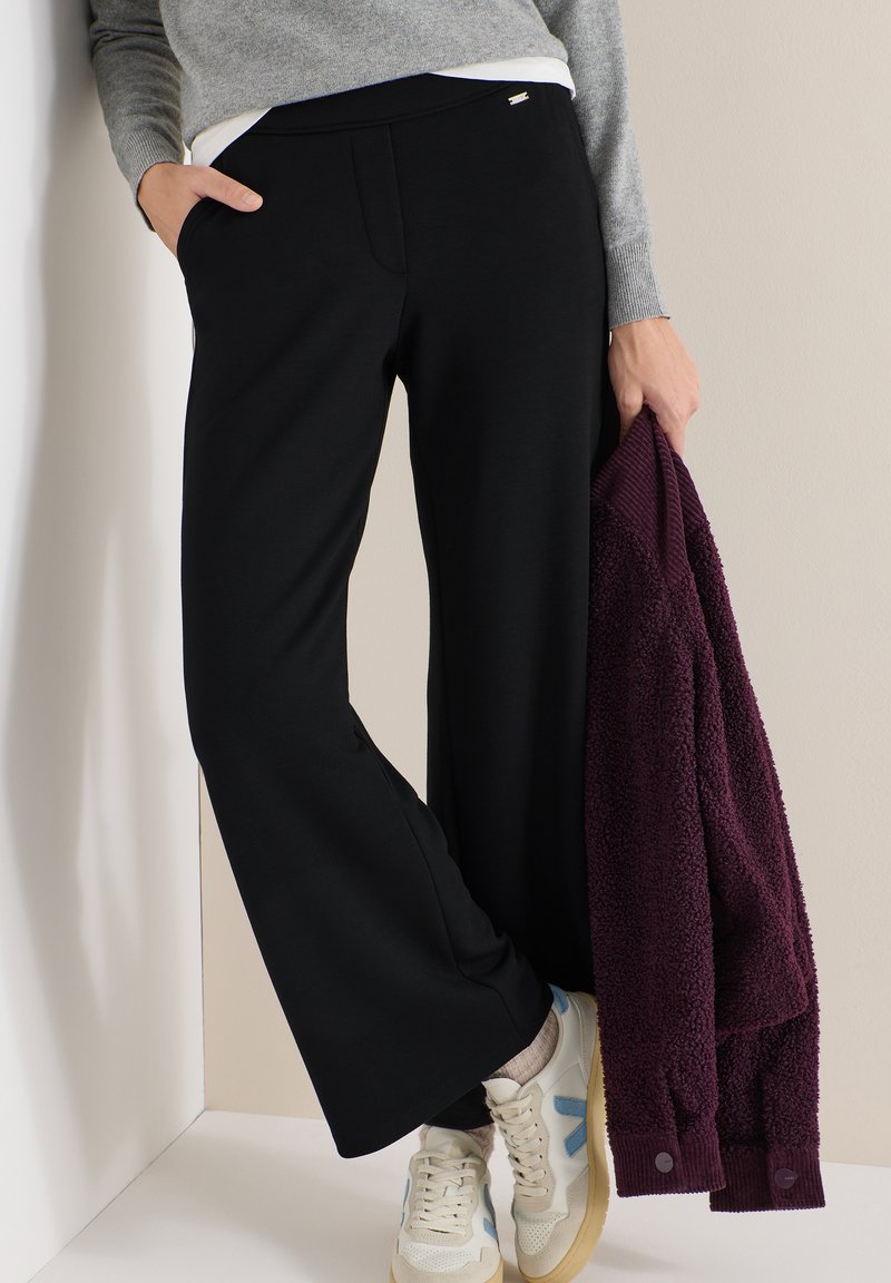 Personne portant un pantalon noir à jambes larges, un pull gris, des baskets blanches, tenant une veste polaire violet foncé pliée dans une main.
