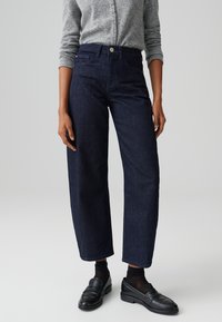 Jeans a gamba larga in denim blu scuro con vita alta, cinque tasche e chiusura con bottone, abbinati a mocassini neri e calze.