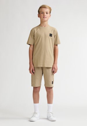 Beige katoenen t-shirt en short set, met een zwart logopatch op de borst en short, gecombineerd met witte sneakers en sokken.