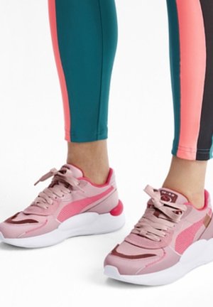 Osoba nosząca różowo-białe trampki Puma w połączeniu z dopasowanymi sportowymi legginsami w kolorach turkusowym i różowym.