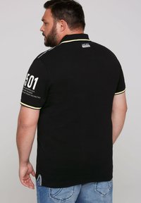 Black polo shirt with yellow and white accents, short sleeves, and a graphic print on the back. Features a label at the collar and side detail.