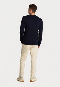 Un uomo sta in piedi di spalle, indossando una maglietta a maniche lunghe blu navy, pantaloni beige, calzini bianchi e sneaker color crema.