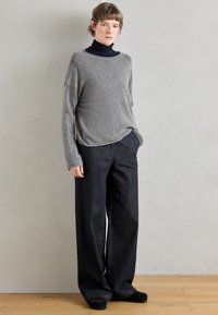 Jasnoszary wełniany sweter o szerokim dekolcie, zestawiony z ciemnoniebieskimi, szerokimi spodniami dżinsowymi. Model stoi na tle o neutralnym kolorze.