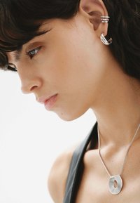Collar de plata con pendant de forma ovalada texturizada, acompañado de múltiples aros de plata, exhibido en un modelo con cabello oscuro.