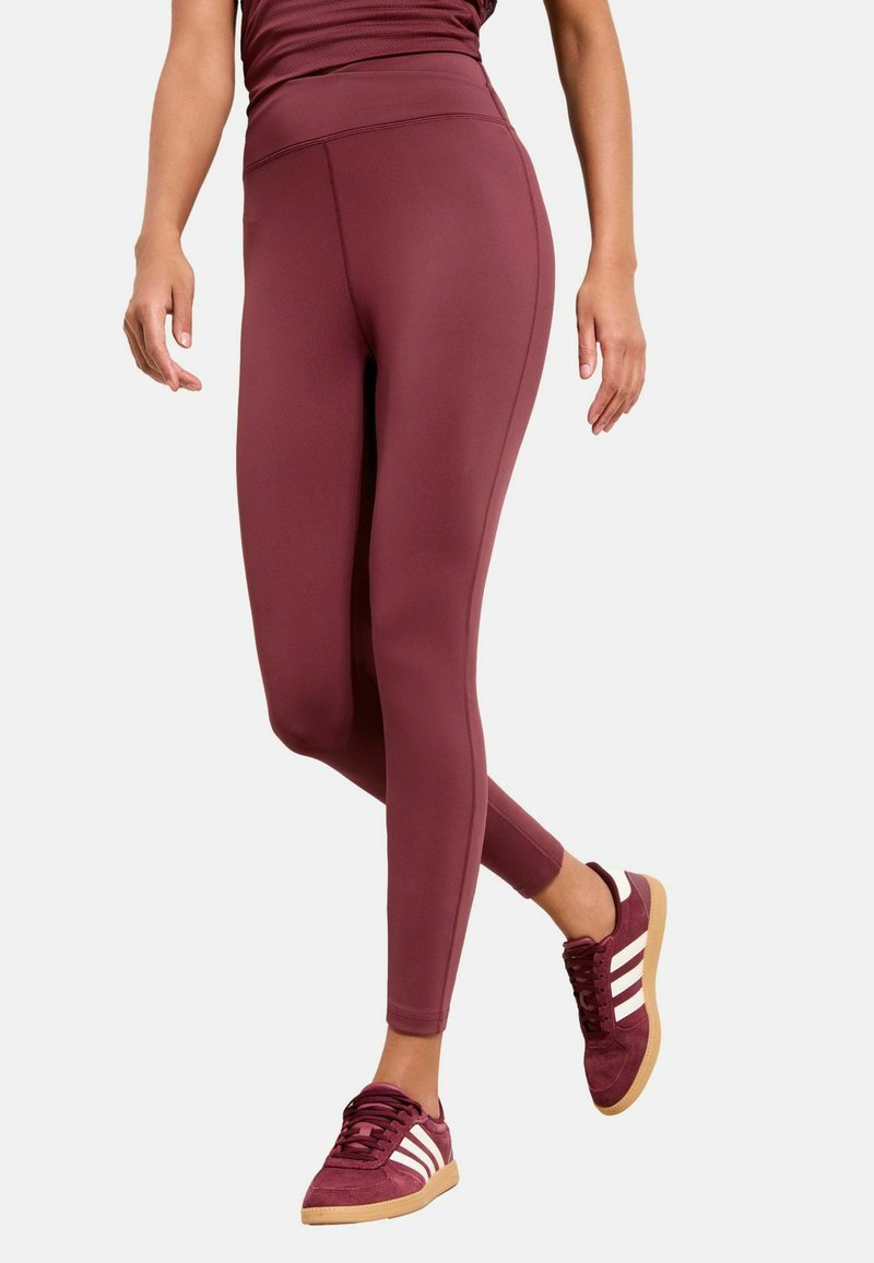 Bordeauxfarbene, aktive Leggings mit hohem Bund, glatter Textur und eng anliegendem Design. Kombiniert mit bordeauxfarbenen Sneakers mit weißen Streifen.