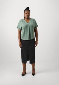 Zeleni, strukturiran vrh z v-neck izrezom in zvitimi rokavi, v kombinaciji s črno krilo do kolen, ki ima sprednji razporek, ter črnimi konicami z visokim petami.