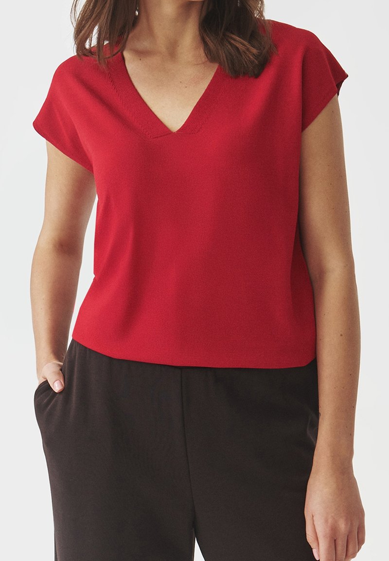 Femme portant un haut rouge à manches courtes en V et un pantalon noir, une main dans la poche sur un fond uni.