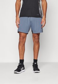 Graue Sportshorts aus synthetischem Stoff, mit einem schwarzen Seitenpanel, akzentuiert durch ein rotes Logo. Getragen mit schwarzen Turnschuhen und Socken.