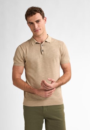Polo bej cu mânecă scurtă, având o textură fină, cu trei nasturi la gât și manșete ribbate, asortat cu pantaloni verzi.