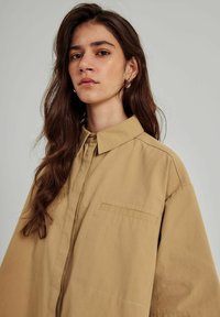 Beige oversized skjorte lavet af bomuld med spids krave, enkelt brystlomme og knaplukning foran. Blød, glat tekstur.