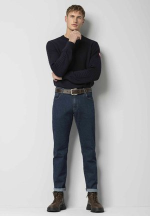 Marineblauwe trui met geribbelde structuur, gecombineerd met donkerblauwe jeans en bruine leren veterschoenen. Model staat tegen een neutrale achtergrond.