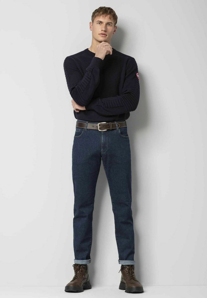 Marineblauwe trui met geribbelde structuur, gecombineerd met donkerblauwe jeans en bruine leren veterschoenen. Model staat tegen een neutrale achtergrond.