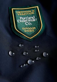 Granatowy materiał z kroplami wody i haftowaną zieloną naszywką w kształcie tarczy z napisem "Portland Outerwear Co." oraz "Outdoor Goods" w białym i złotym kolorze.